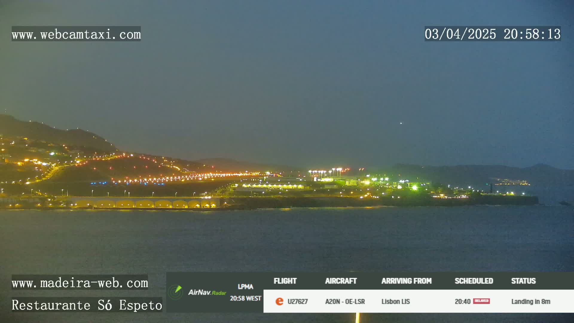 A nighttime coastal scene shows a brightly lit airport and city on a hillside overlooking dark water under a twilight sky.