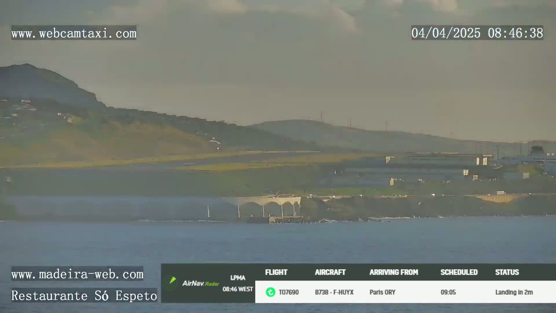 A hazy, overcast day shows an airport runway, coastal buildings, and calm ocean water near a hill.