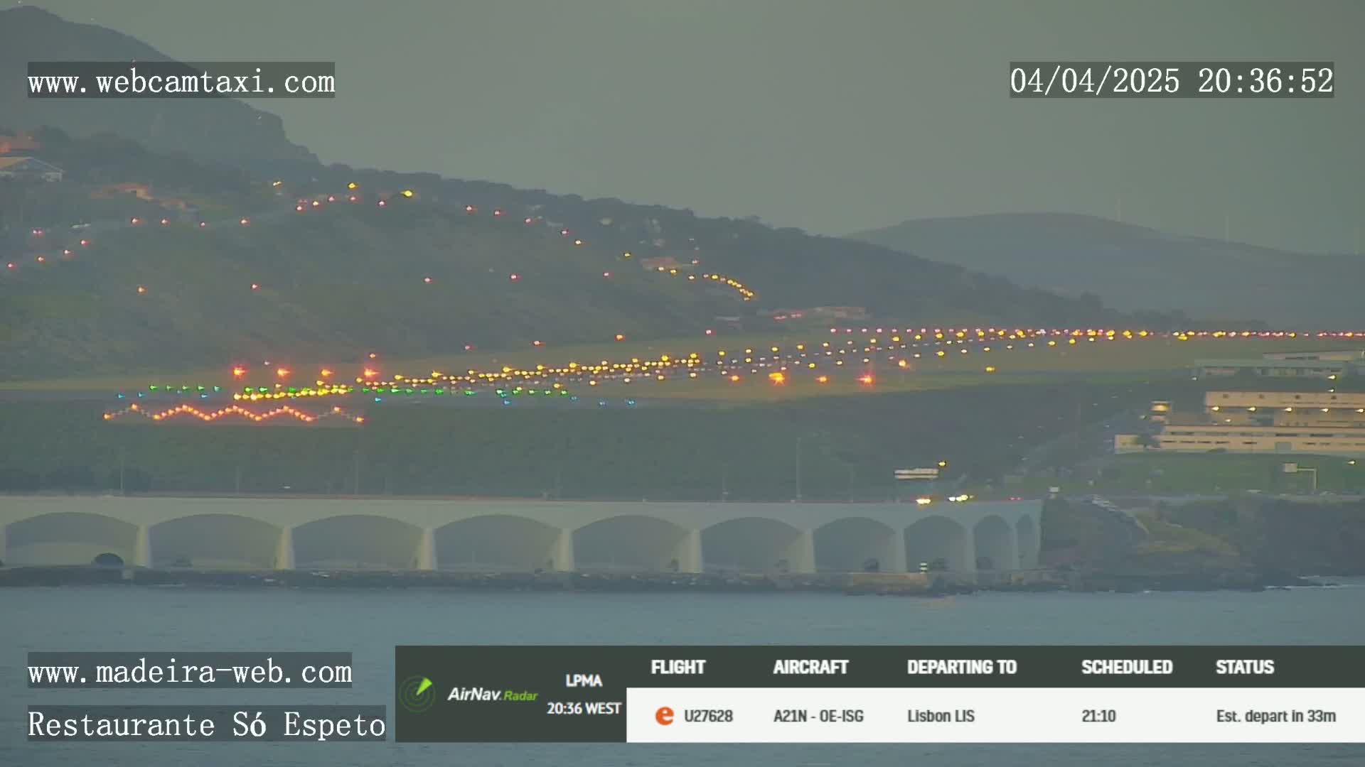A dimly lit airport runway with many lights is visible across a body of water under an overcast sky.