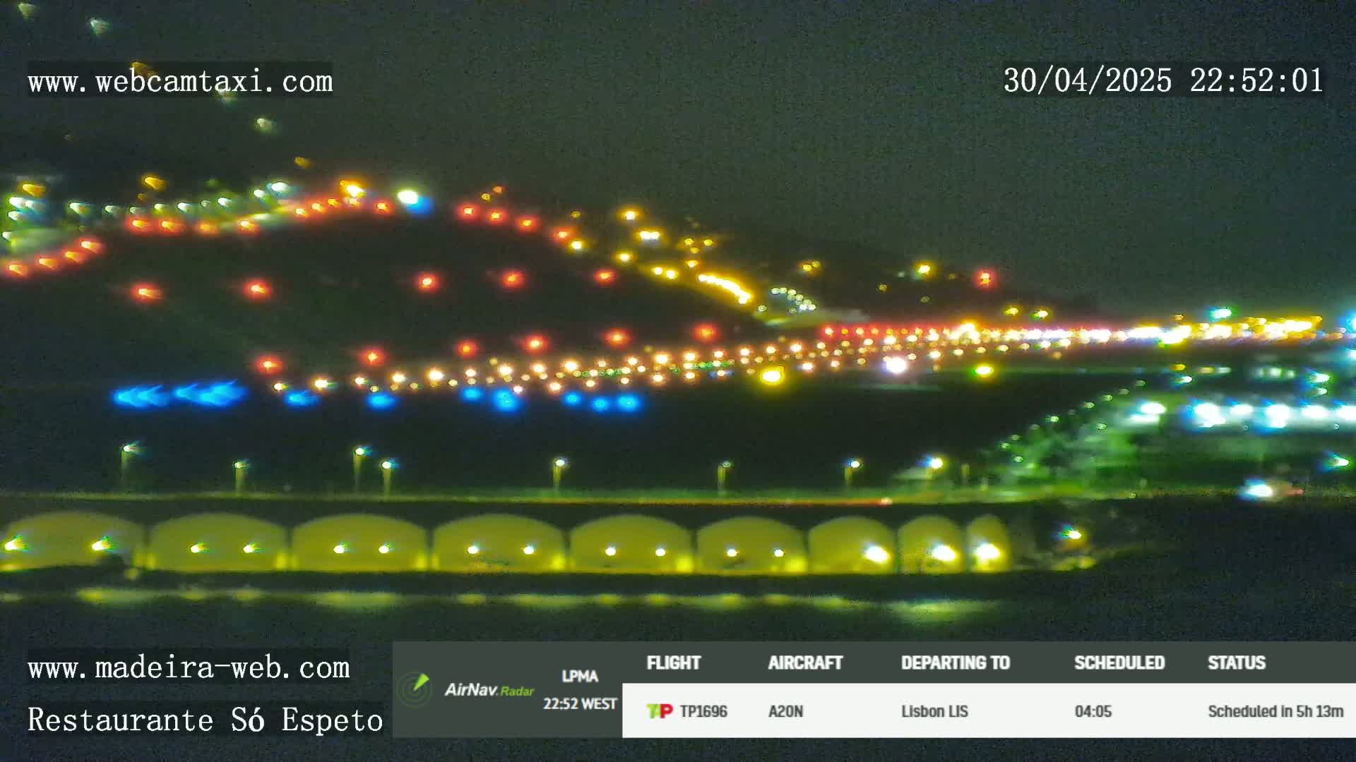 At night, numerous lights illuminate a hilly landscape near a body of water, with a bridge and more lights visible in the distance.
