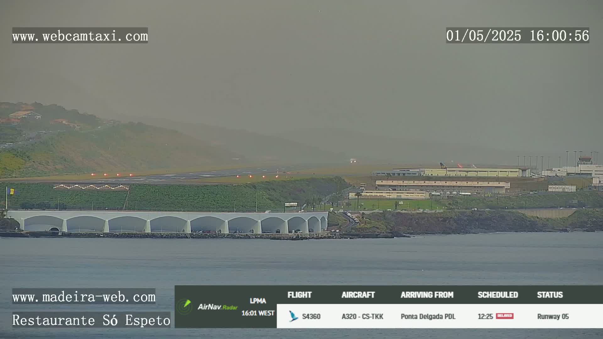 A hazy view shows an airplane landing on a runway near a coastline, with a long, low building along the water's edge.
