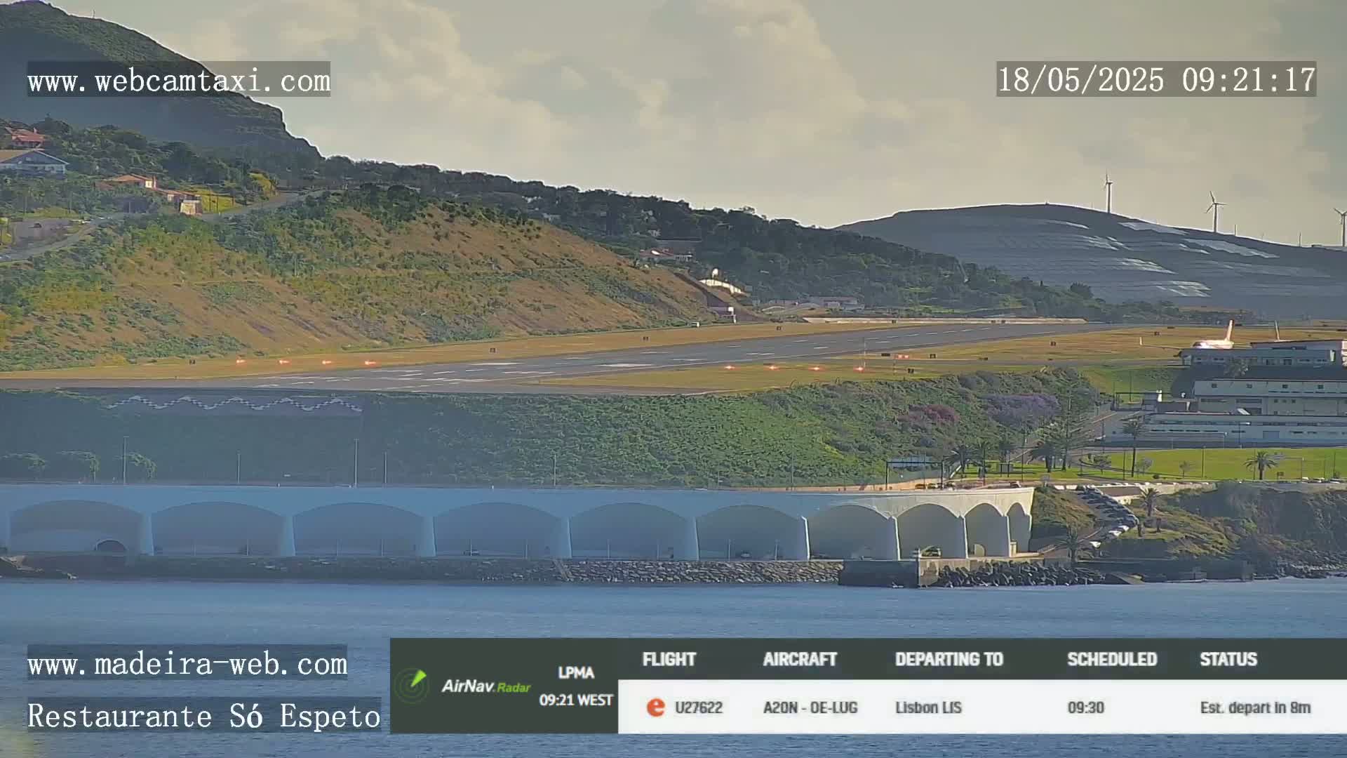 An airport runway with a plane visible near the terminal, nestled between a hillside and the ocean under mostly cloudy skies.