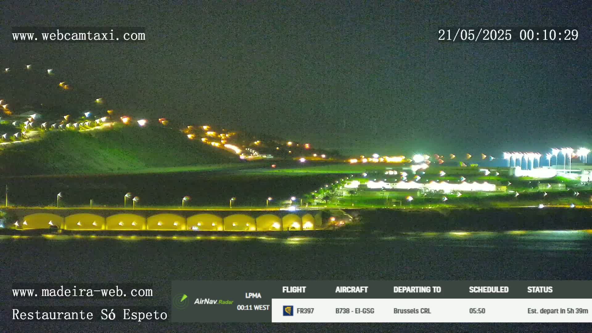 A nighttime, seaside view shows a hill with lights, a long structure with arches, and a distant airport with bright lights under a clear, dark sky.
