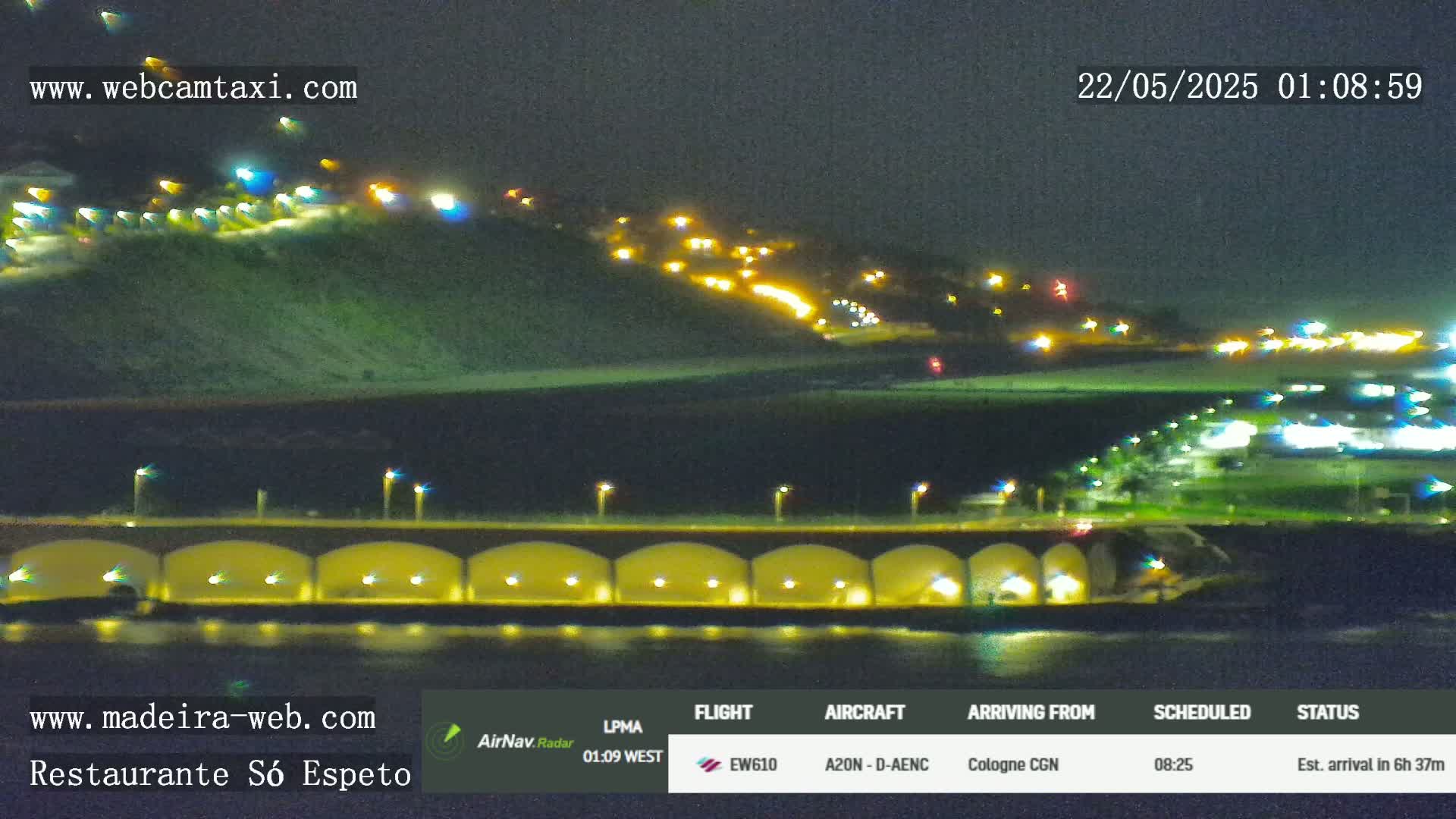 A nighttime coastal scene shows a hilltop with scattered lights, a bridge with illuminated arches over dark water, and more lights in the distance.