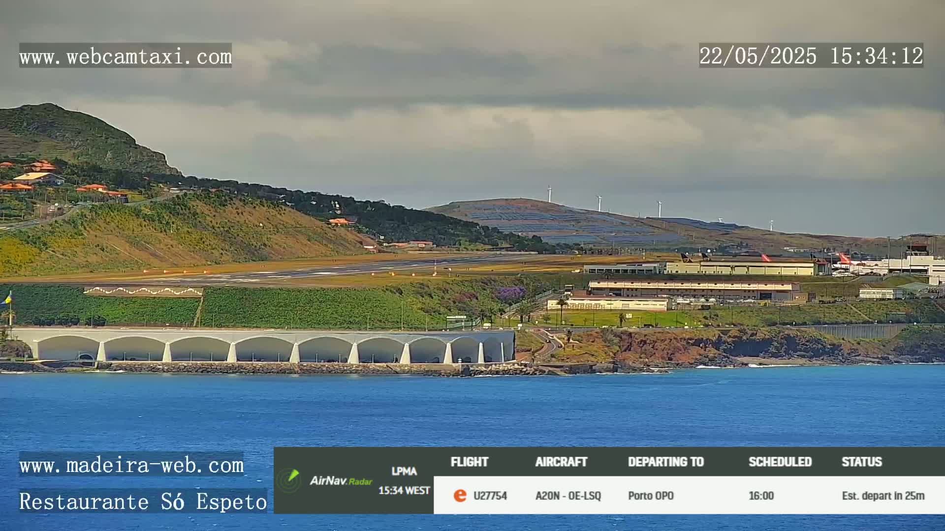 An airport runway and terminal are visible near a calm ocean under a cloudy sky, with a hillside and wind turbines in the background.