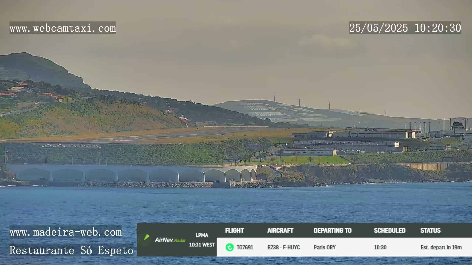 An airport runway and terminal are situated on a coastal hillside overlooking calm ocean waters under a hazy sky.