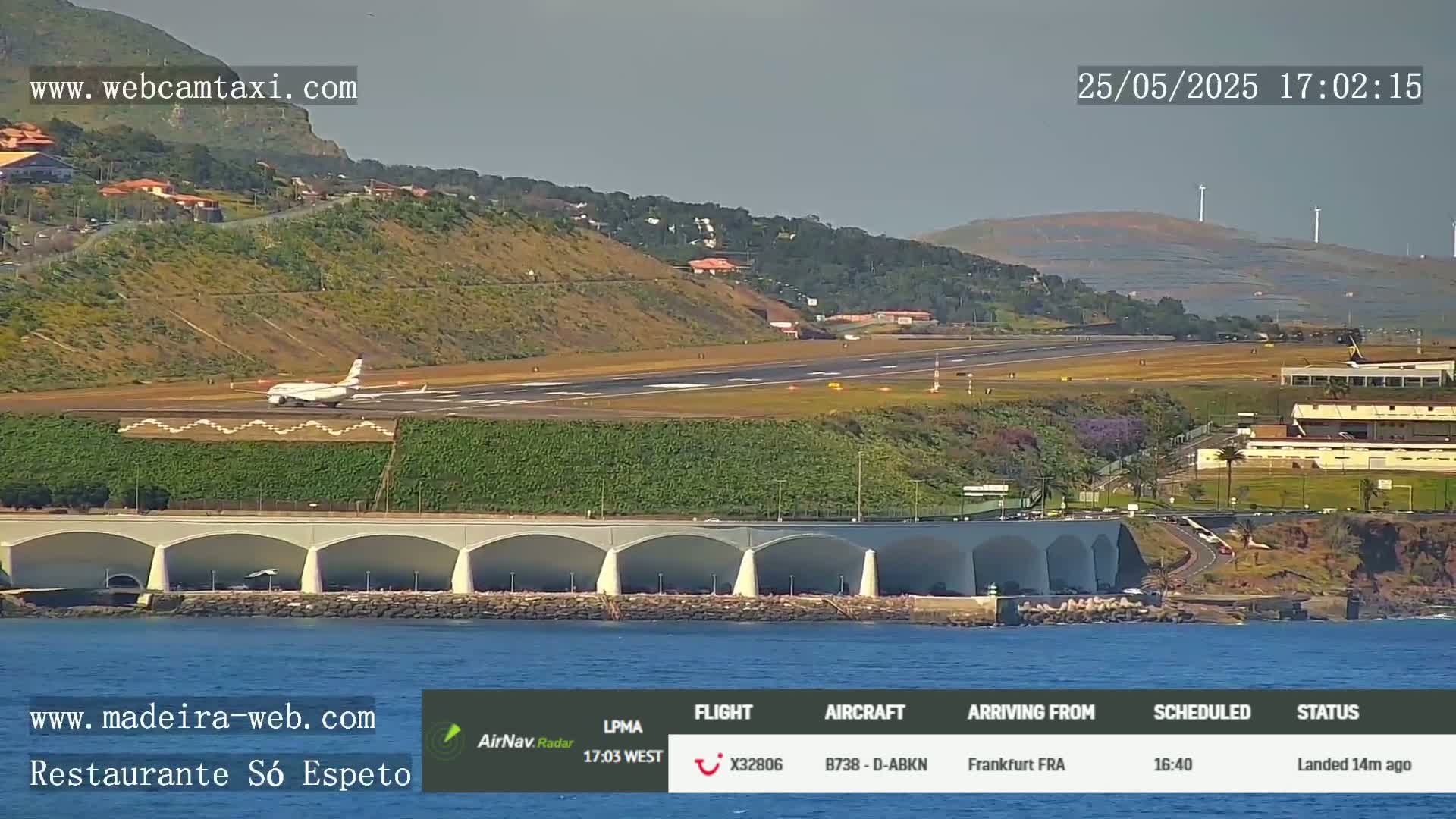An airplane is landing on a runway near a coastal area with calm water and a curved wall structure, under a mostly clear sky.