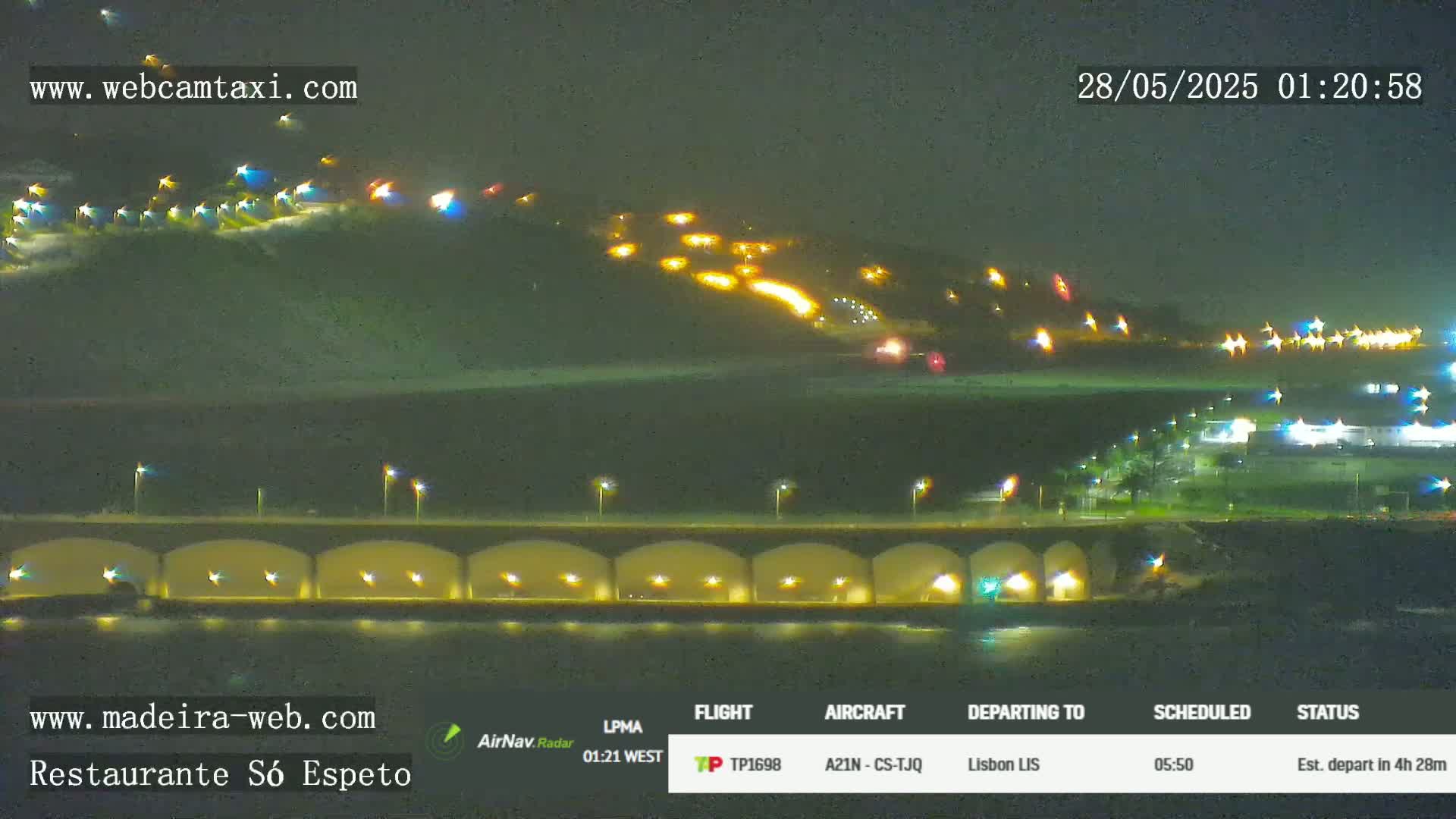A nighttime long shot shows a hilltop with numerous lights, a bridge with lights under it, and a body of water, all under a dark sky.