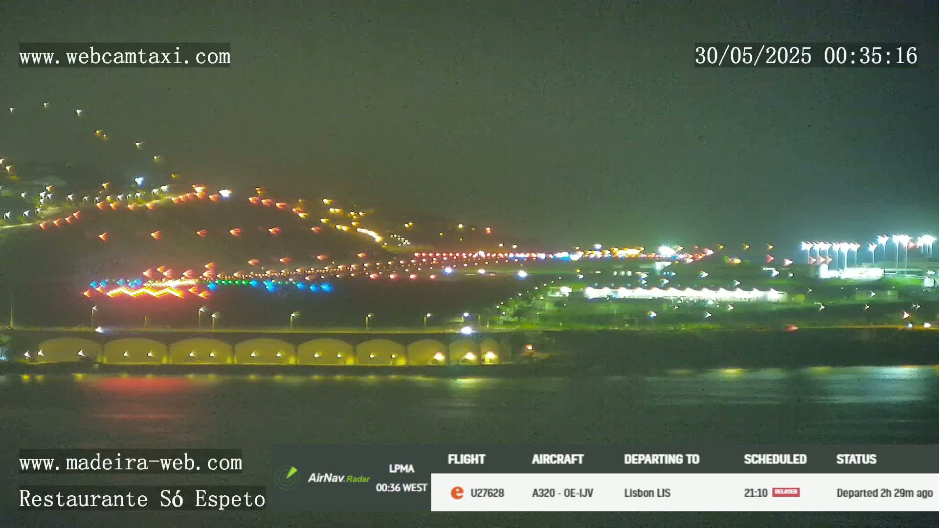 A nighttime long shot of an airport runway with numerous lights, adjacent to a body of water under a dark sky.