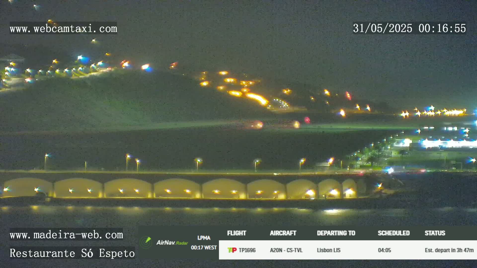 A nighttime long shot shows a hillside with lights, a bridge with lights under it, and a body of dark water.