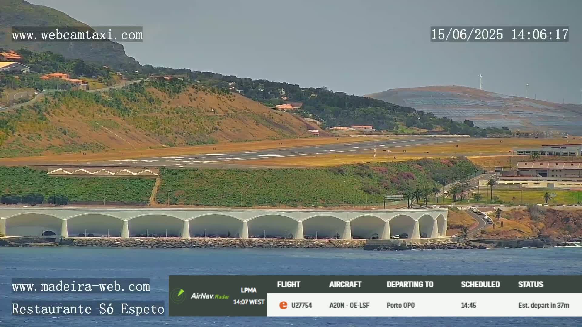 An airport runway and terminal are visible near a coastline under a clear sky, with a structure featuring arched alcoves along the water's edge.