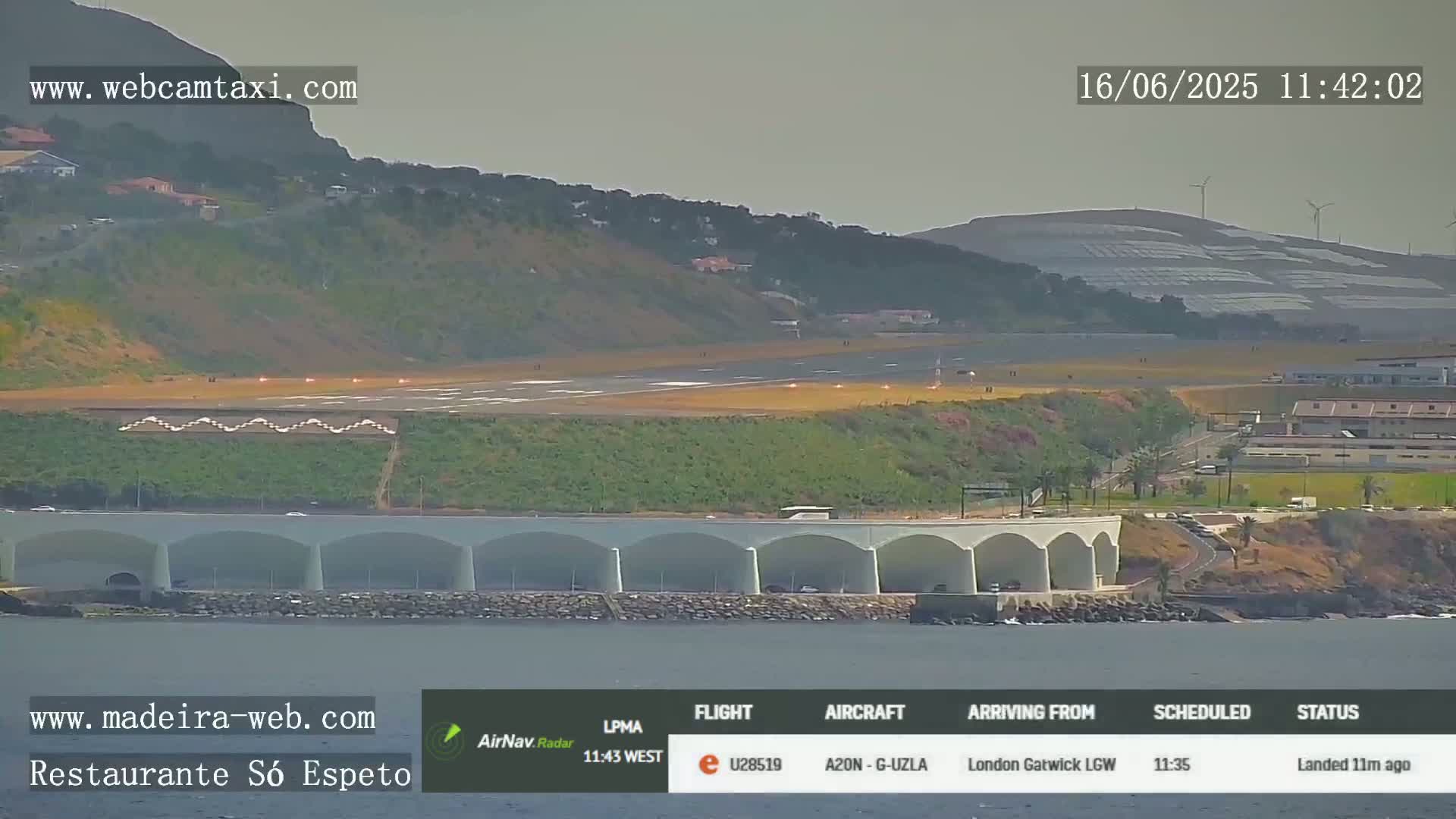 A long, flat runway is visible at an airport near the ocean, with a structure resembling a bridge or overpass nearby, under a hazy sky.
