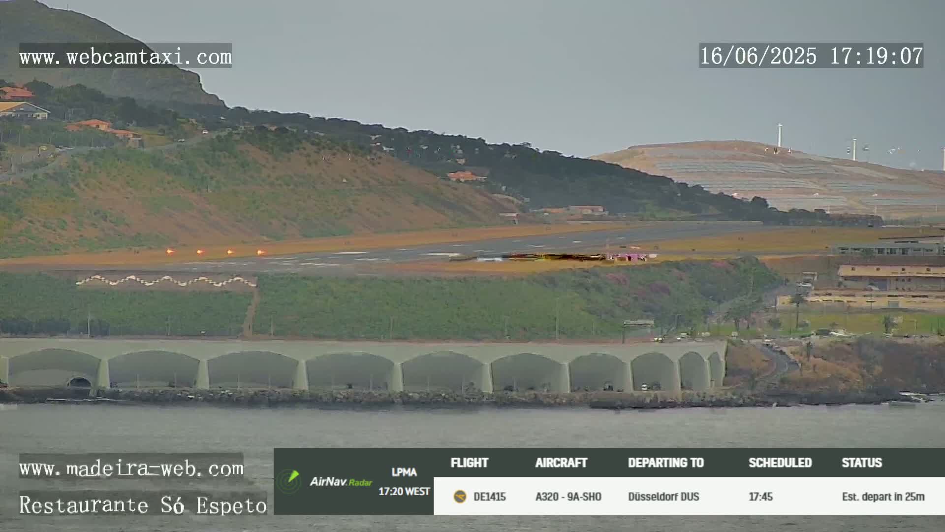 A long, low building with arched openings sits by the ocean next to an airport runway where a plane is preparing for takeoff under an overcast sky.