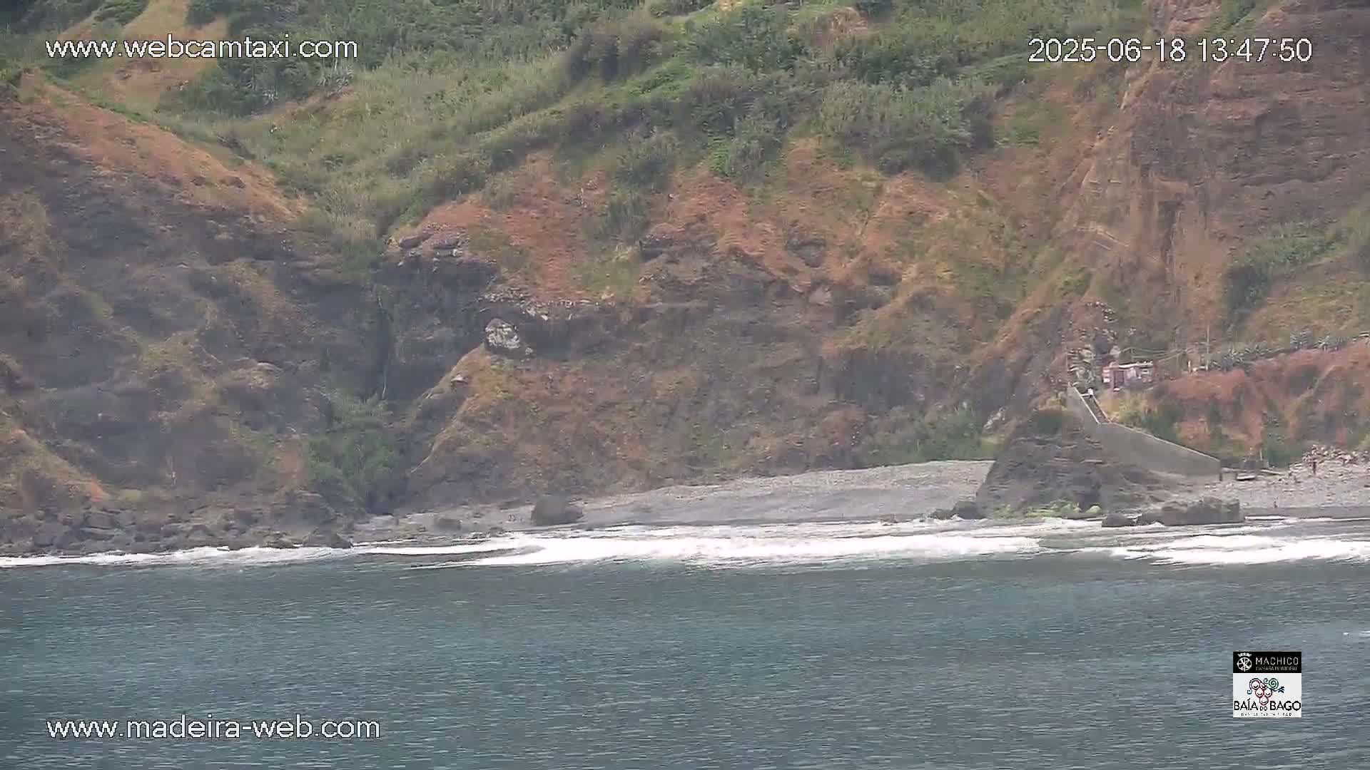 A dark, rocky cliff overlooks a small, dark-sand beach with gentle waves lapping the shore.