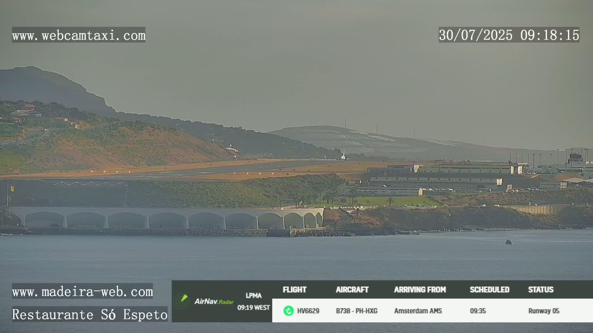 A distant airplane approaches a runway near a body of water under hazy, light conditions.