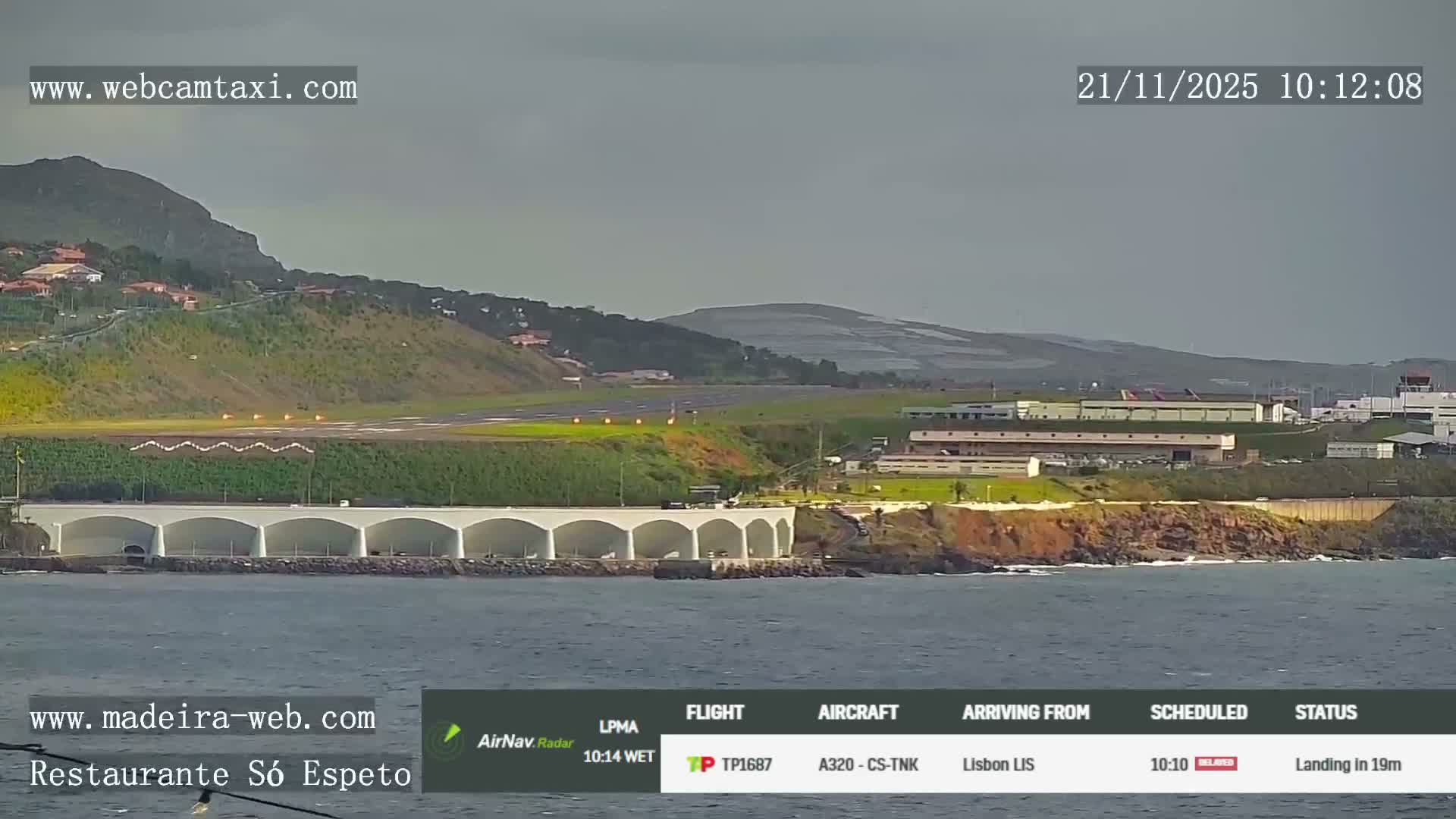 An expansive coastal airport runway extends over an arched pier into the sea, with green hills and scattered buildings in the background under an overcast sky.