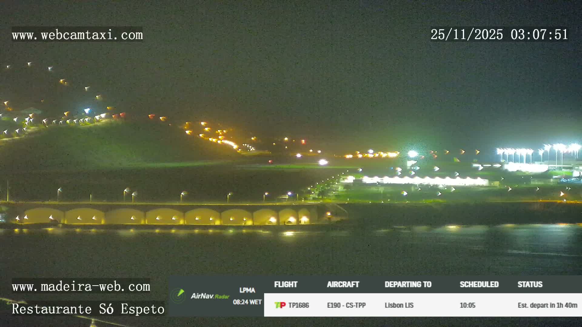 A clear night view reveals an illuminated airport facility next to a body of water, featuring a long arched bridge and numerous scattered lights across a distant hillside.