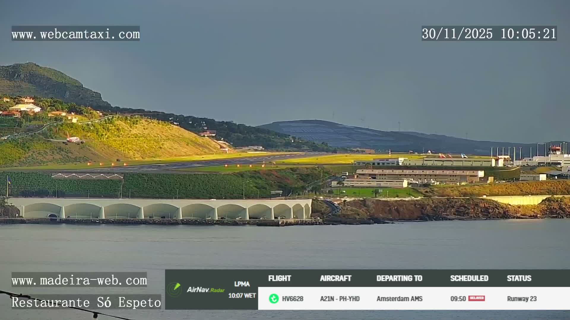 A commercial airplane is visible on the long runway of a coastal airport, which extends over the sea supported by a multi-arched viaduct, all nestled among green hills under an overcast sky.