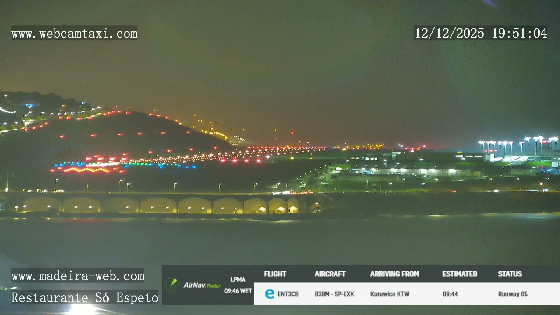 A commercial airplane is visible on the long runway of a coastal airport, which extends over the sea supported by a multi-arched viaduct, all nestled among green hills under an overcast sky.