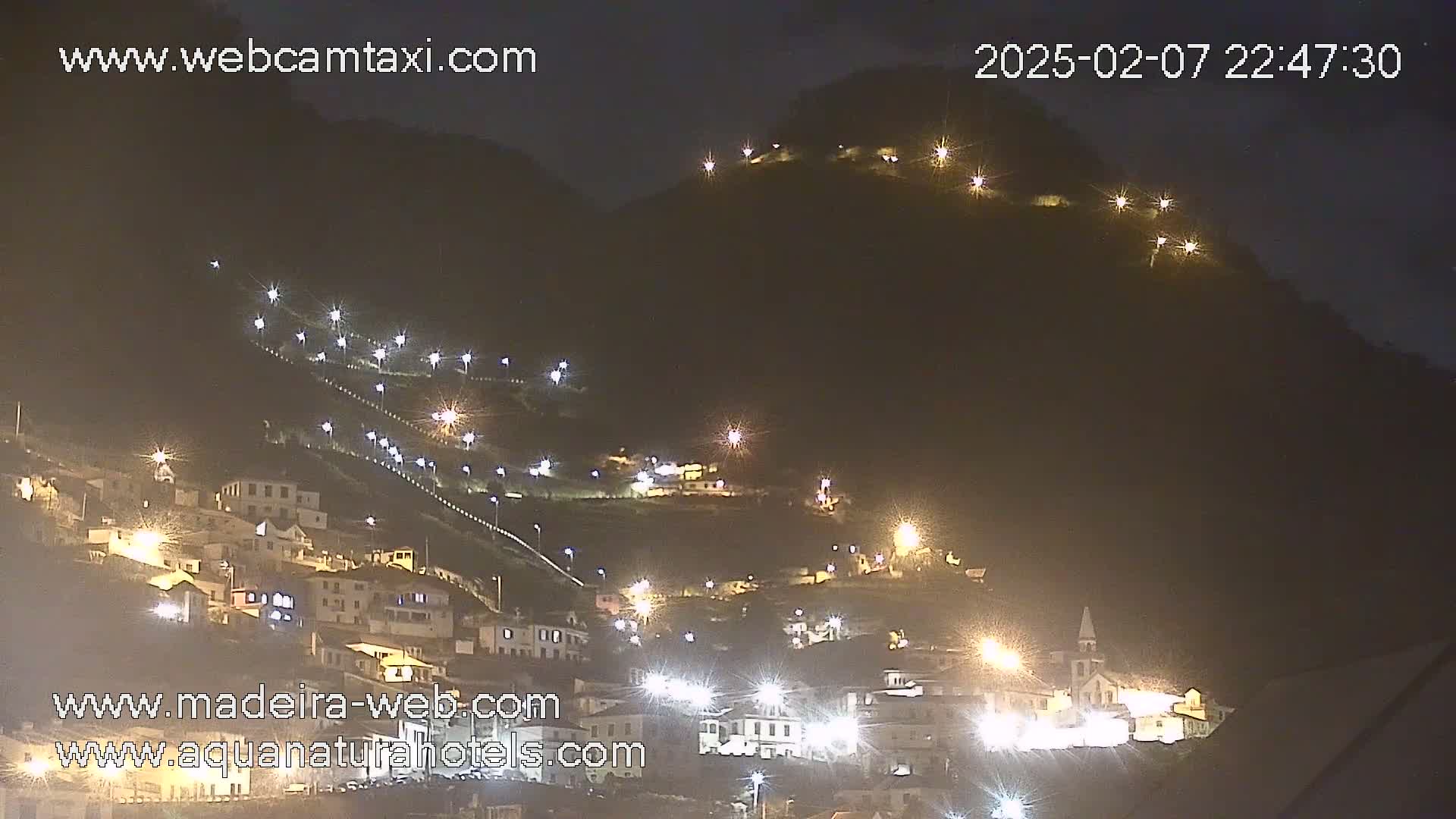 A nighttime view of a hillside town illuminated by streetlights, under a clear, dark sky.
