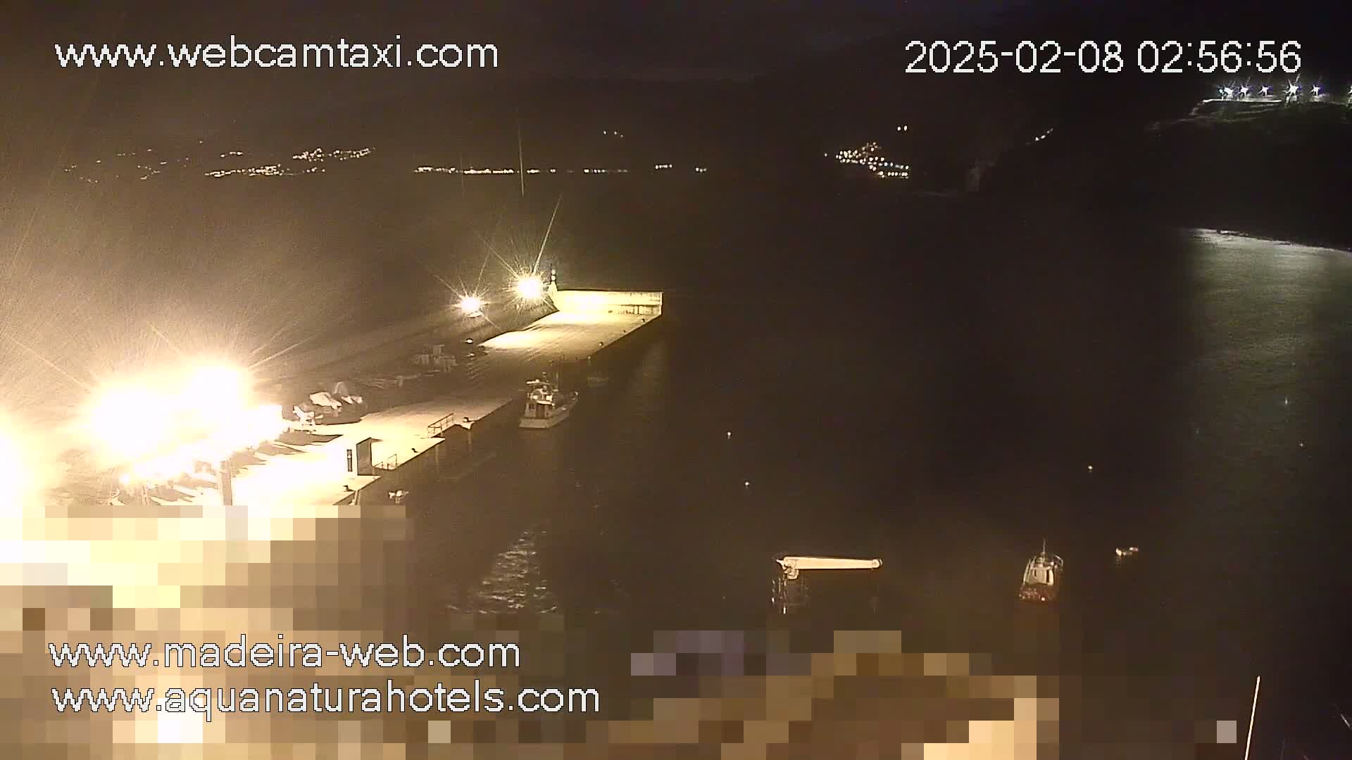 A nighttime view of a harbor with several boats moored at a brightly lit pier, under a calm, dark sky.