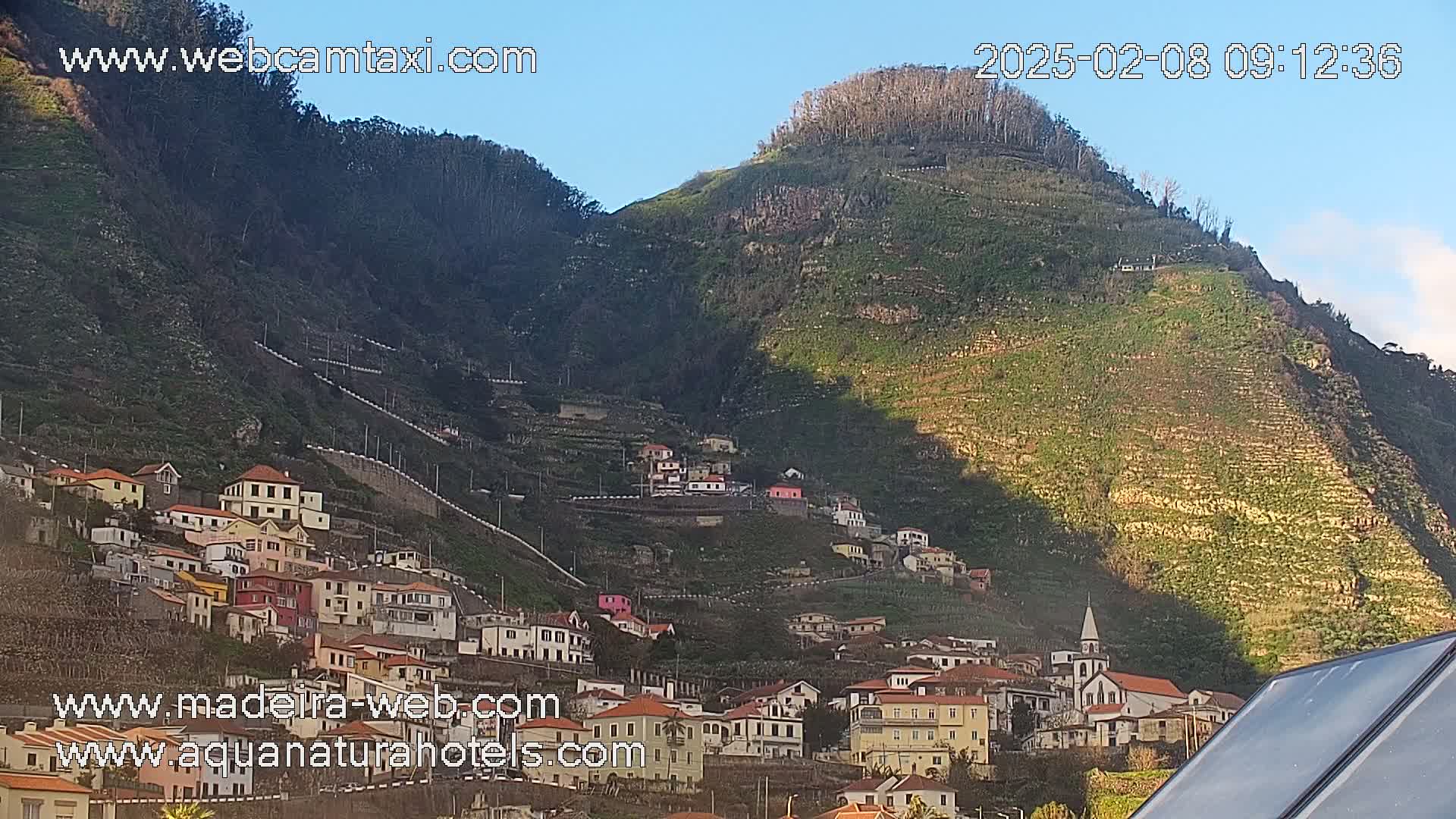 A hillside village with colorful houses clings to the steep, terraced slopes of a green mountain under a clear blue sky.
