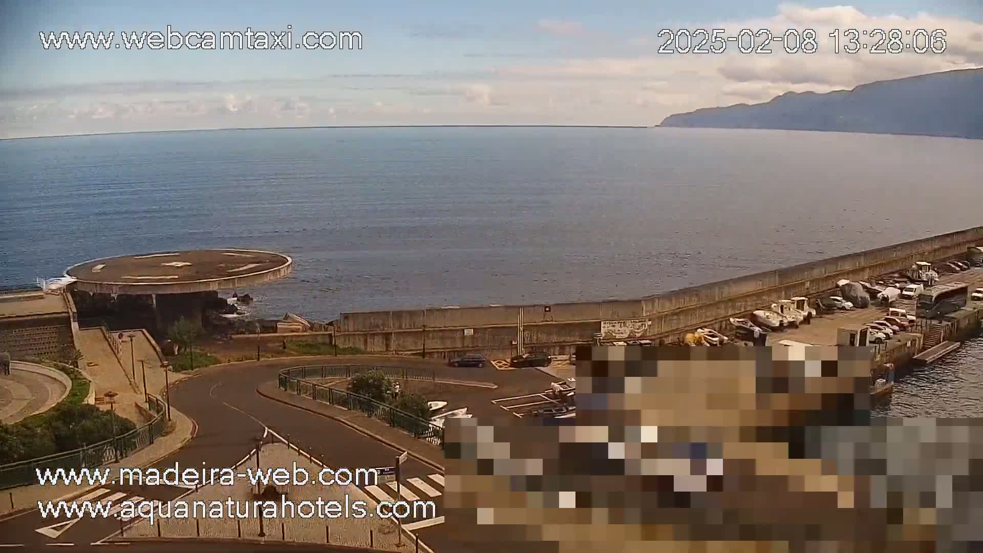 A mostly sunny day overlooks a calm ocean, a circular helipad, a seawall with parked vehicles, and a coastal road curving away from the viewer.