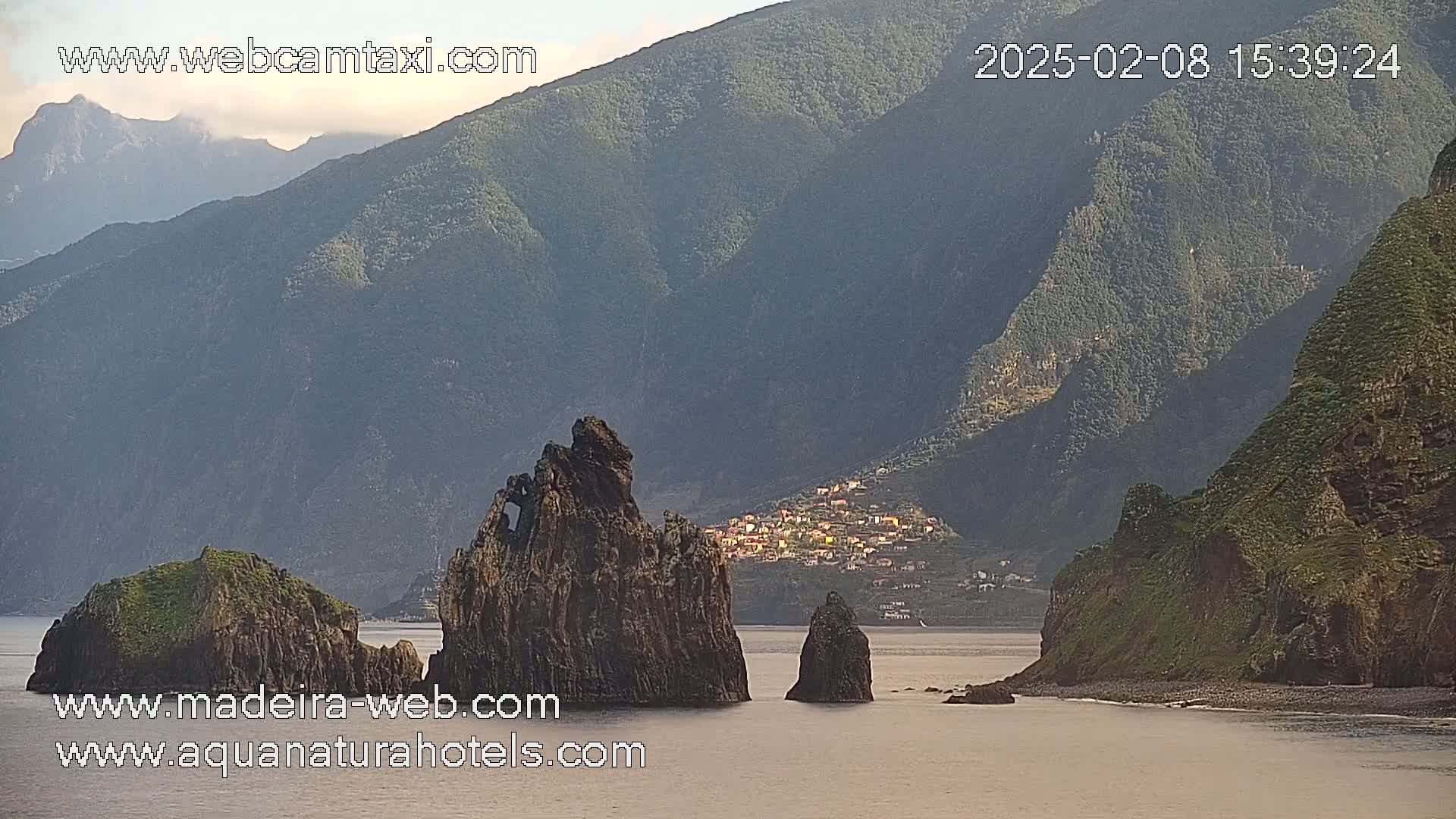 A calm body of water laps against a rocky shoreline, featuring several prominent rock formations in the foreground, with a village nestled in the hills in the background under a partly cloudy sky.