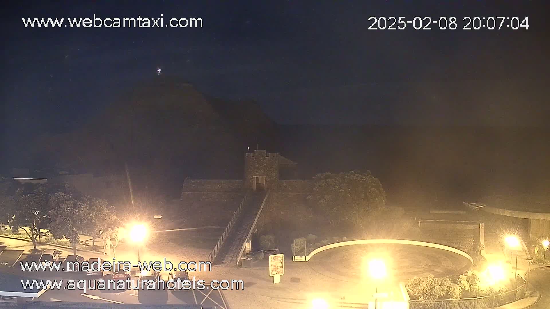 A nighttime, outdoor view shows a stone structure, possibly a fortress or castle, illuminated by artificial lights, with a dark hill in the background under a clear, dark sky.