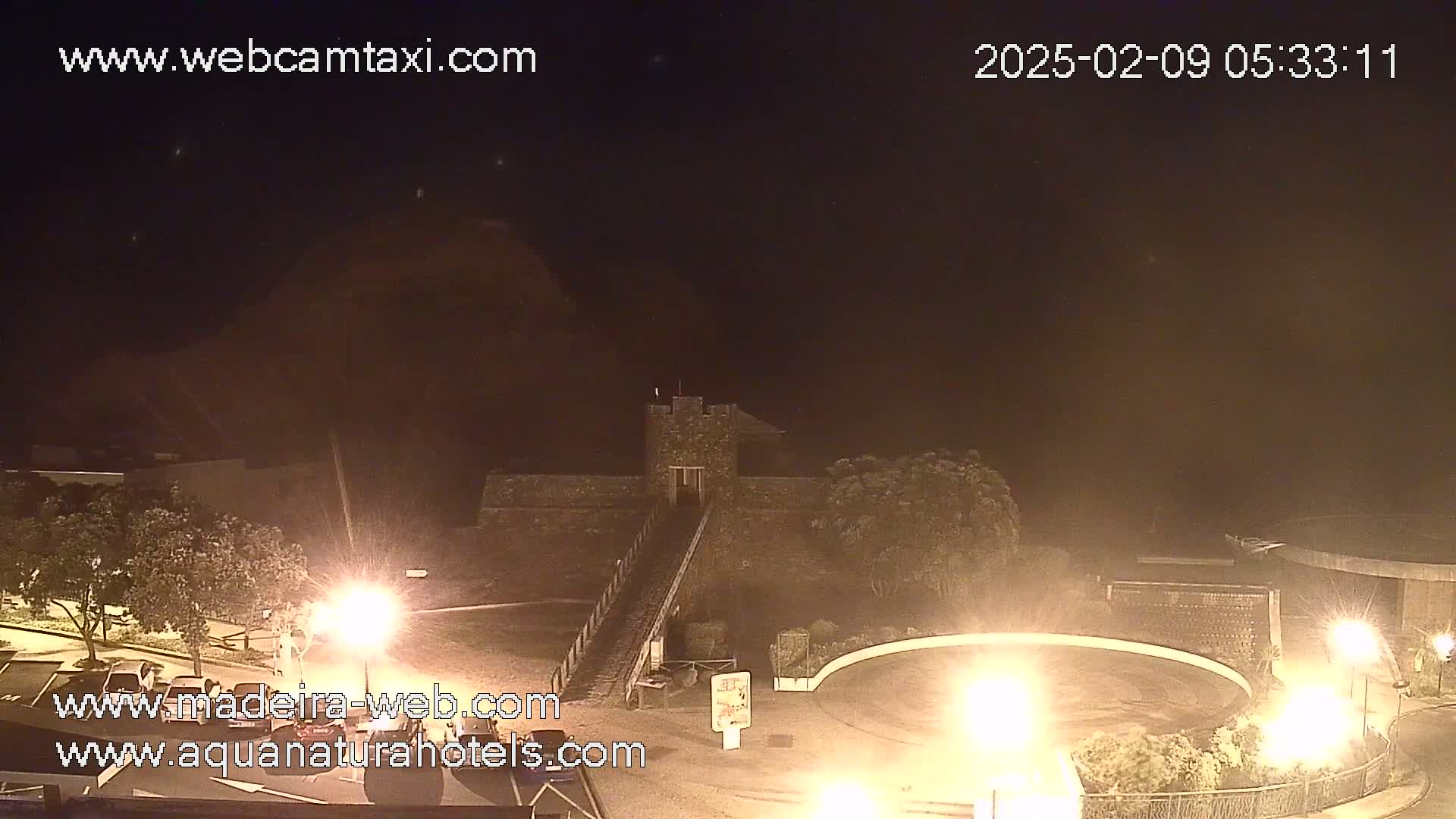 A nighttime view of a stone structure resembling a castle, illuminated by artificial light, with some vegetation and a paved area visible.