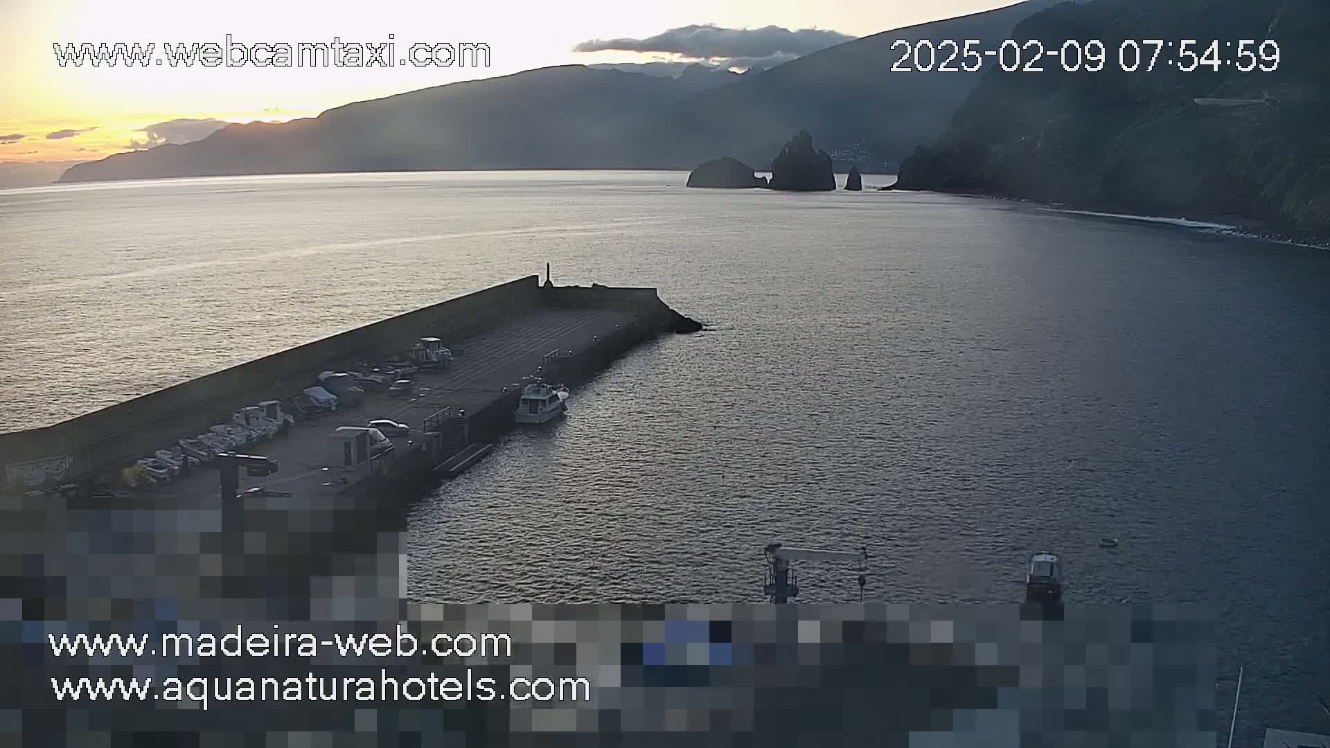 A sunrise illuminates a calm harbor with parked cars and boats near a rocky coastline and distant mountains.