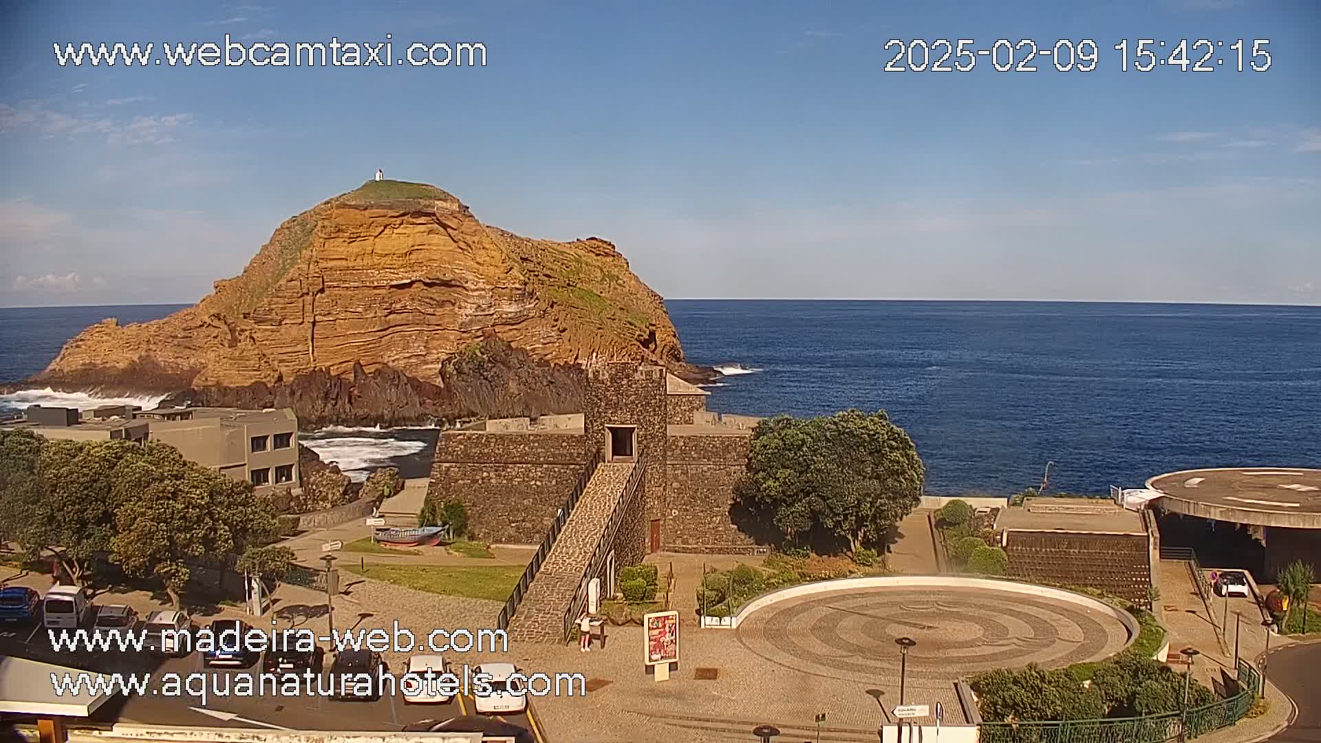 A rocky, reddish-brown island with a small structure on top sits in a calm, blue ocean next to a stone fort and a circular plaza on a sunny day.