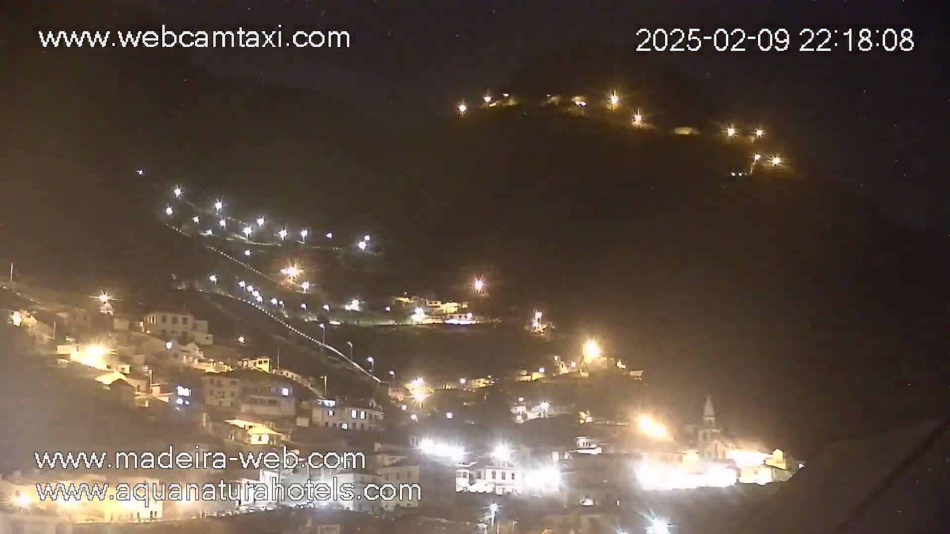 A hillside town is illuminated at night, with many lights visible on the buildings and streets.