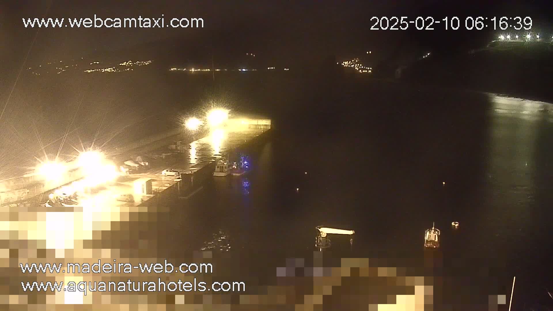 A nighttime, brightly lit harbor scene shows several boats moored at a dock, with calm water extending into the darkness beyond.