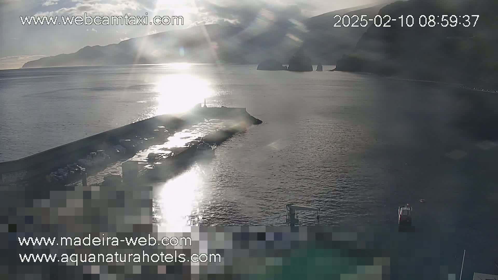 A partly sunny, somewhat hazy day shows a harbor with a long breakwater, boats, and a mountainous coastline in the distance.
