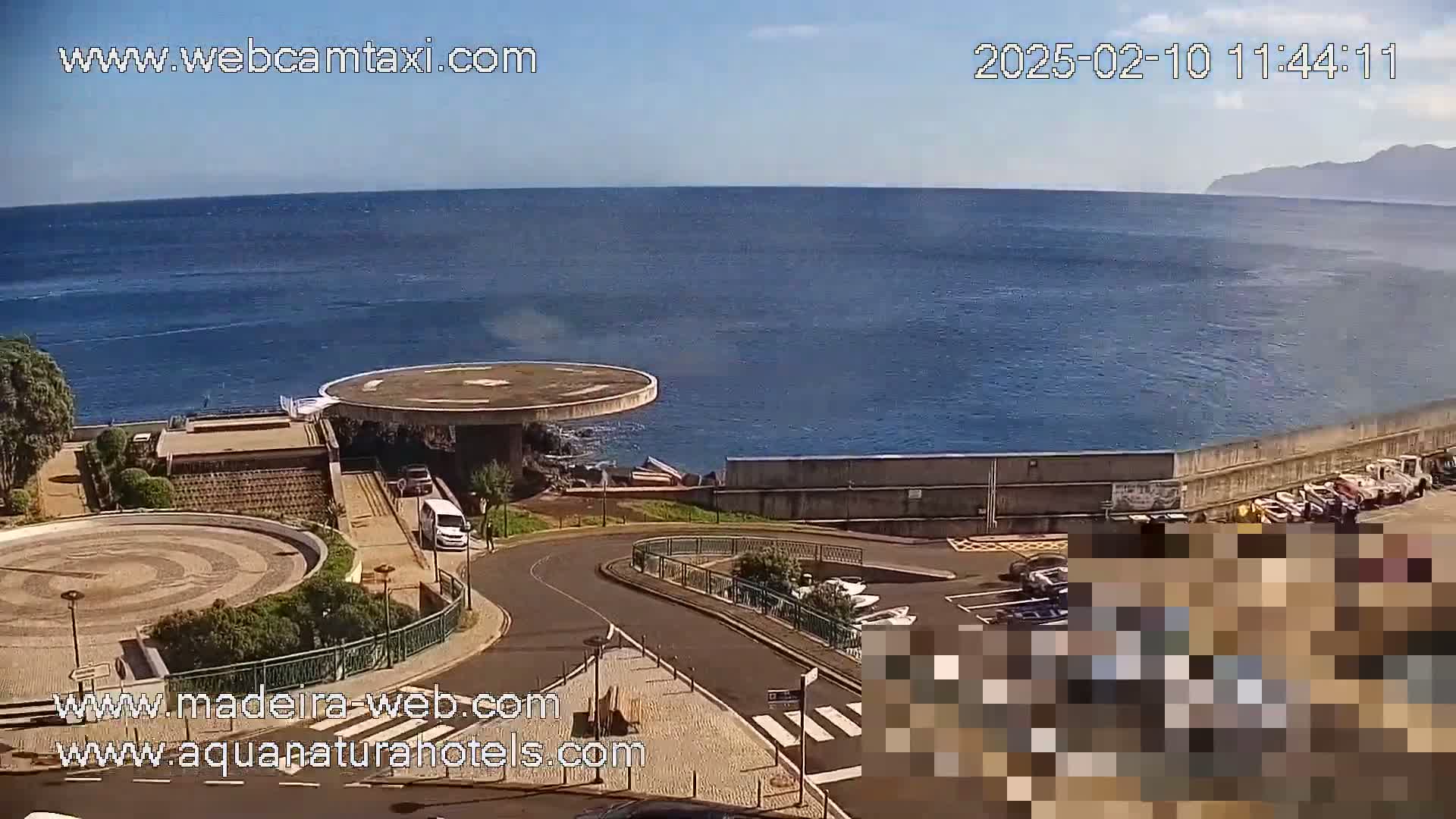 A seaside view on a sunny day shows a circular structure by the water, a paved area with cars parked nearby, and a calm ocean extending to a distant shoreline.