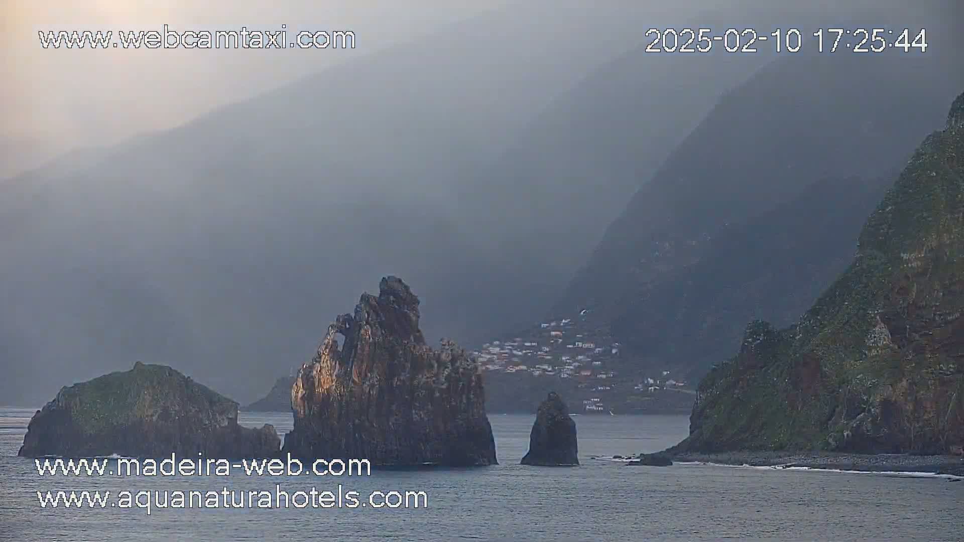 A hazy, overcast scene shows a coastal village nestled between dark, steep cliffs and the ocean, with several prominent rock formations in the foreground.