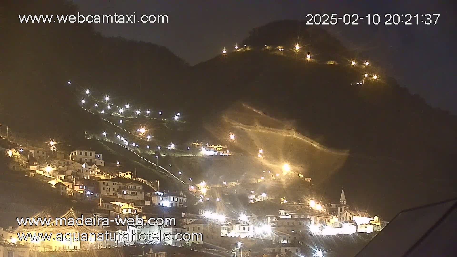 A nighttime view of a hillside town illuminated by streetlights, nestled against a dark mountain under a clear sky.