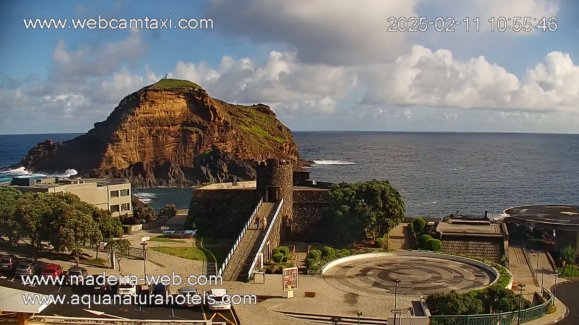 A coastal scene shows a rocky, reddish-brown island off a coastline with a stone structure and bridge, under partly cloudy skies.