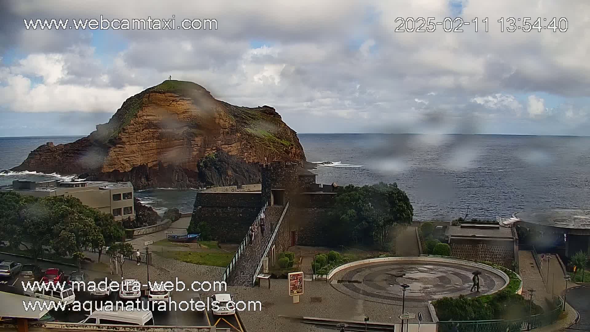 A rain-slicked coastal scene shows a rocky islet with a fortress near a town square, all overlooking a dark ocean under a partly cloudy sky.