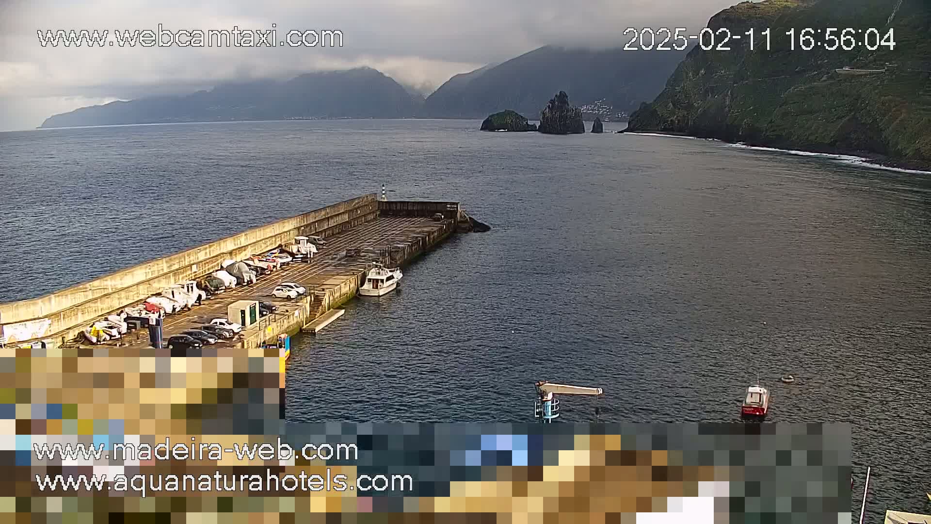 A harbor with several boats and cars parked, under overcast skies, is situated near mountainous terrain.