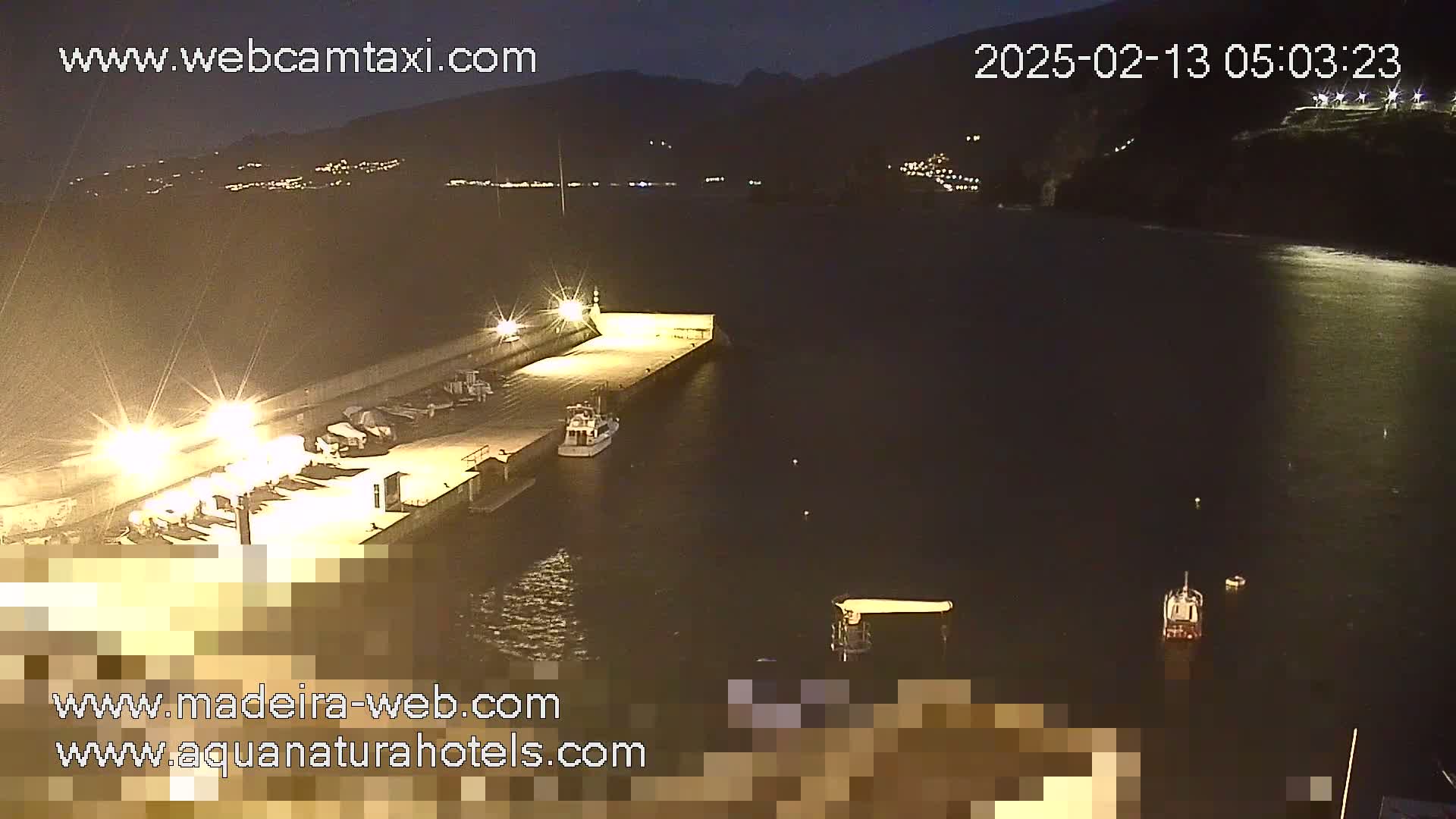 A nighttime view of a harbor with boats docked, illuminated by lights, and a calm body of water extending to a dark hillside in the background.