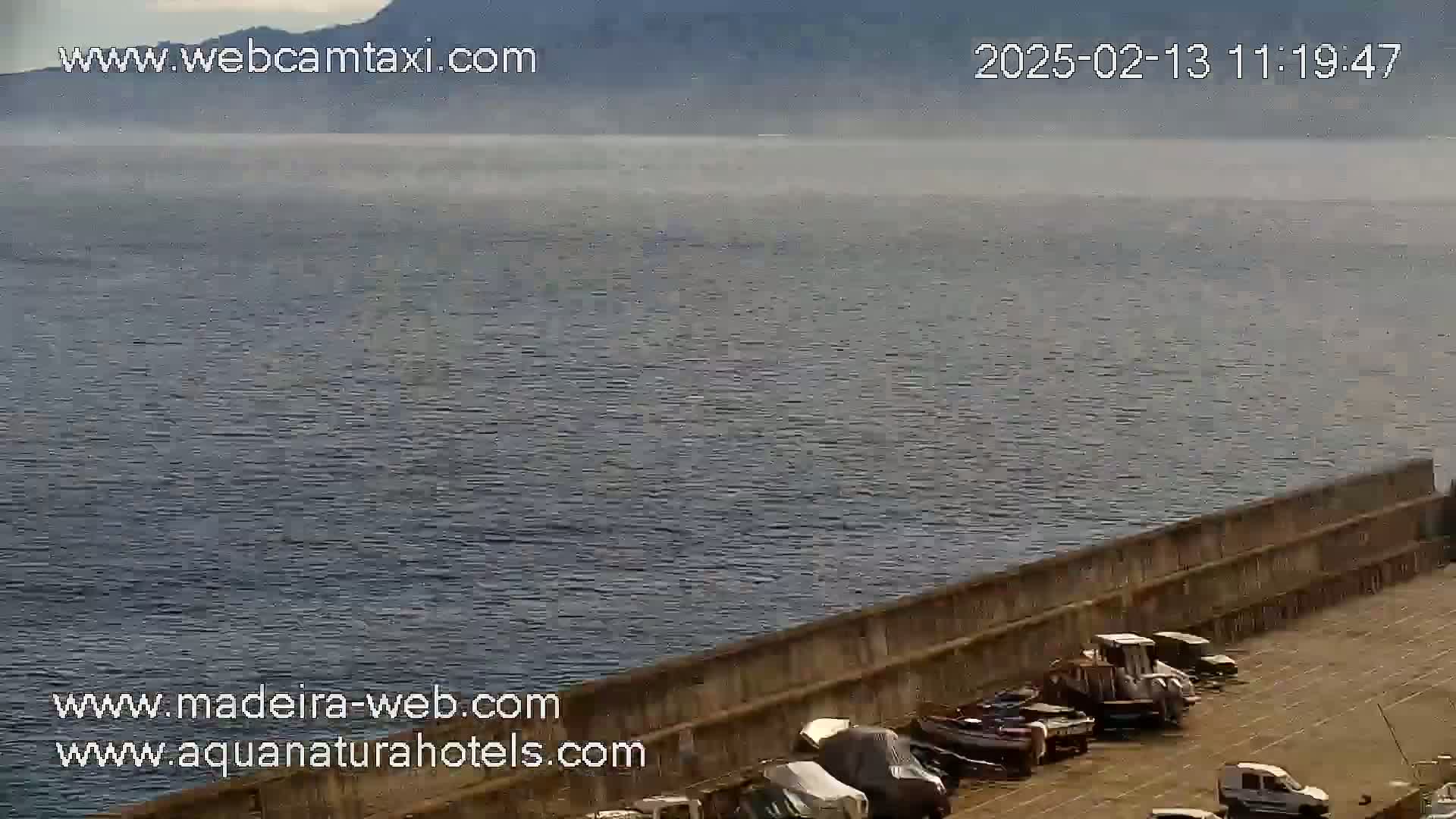 A hazy view of a calm body of water extends to distant mountains, with a seawall and several parked vehicles visible in the foreground.