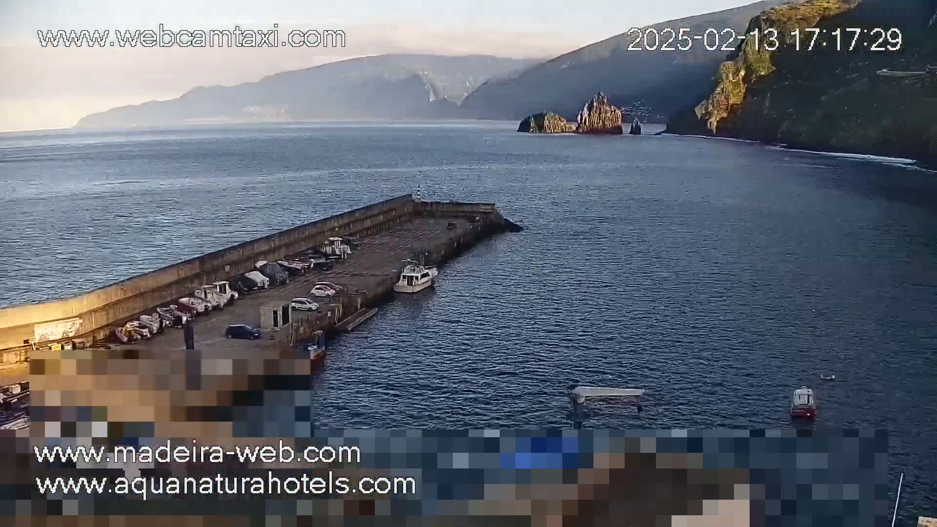 A calm body of water is shown with a long concrete pier containing several parked vehicles and a few small boats, and in the background, high rocky cliffs are visible under a clear sky.