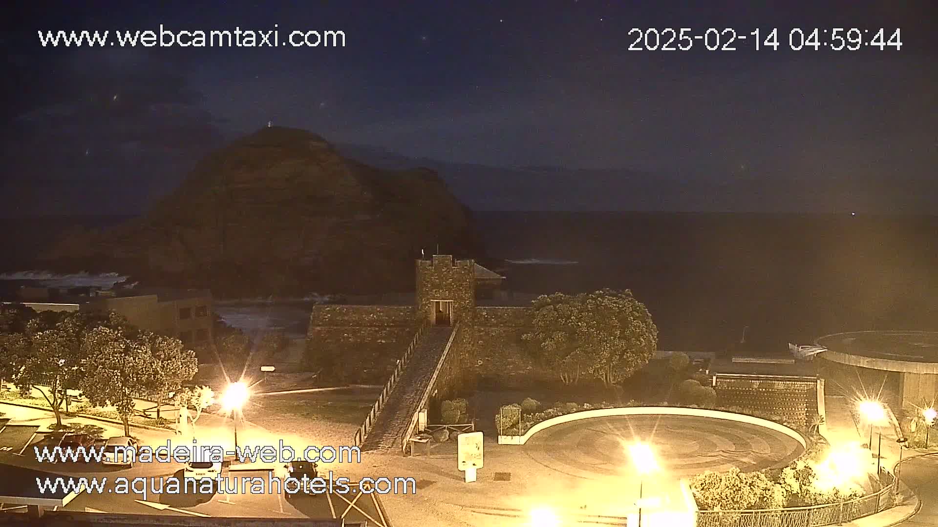 A nighttime coastal scene shows a stone structure with a bridge leading to it, a hill in the background, and a circular roadway lit by streetlights, with a dark ocean visible in the distance and some wave action.