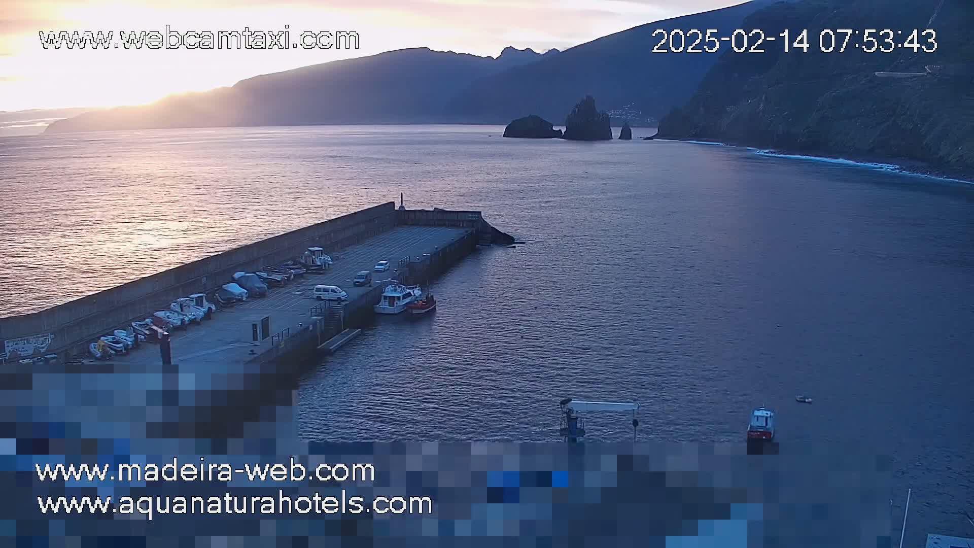 A calm body of water at sunset is viewed from above, showing a small harbor with several boats and cars parked along a concrete wall, with dark mountains in the background.