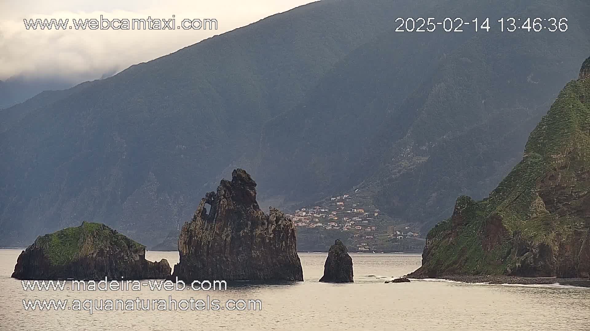 A hazy, overcast day reveals several dark, rocky outcroppings in calm ocean water near a village nestled on a steep, green hillside.