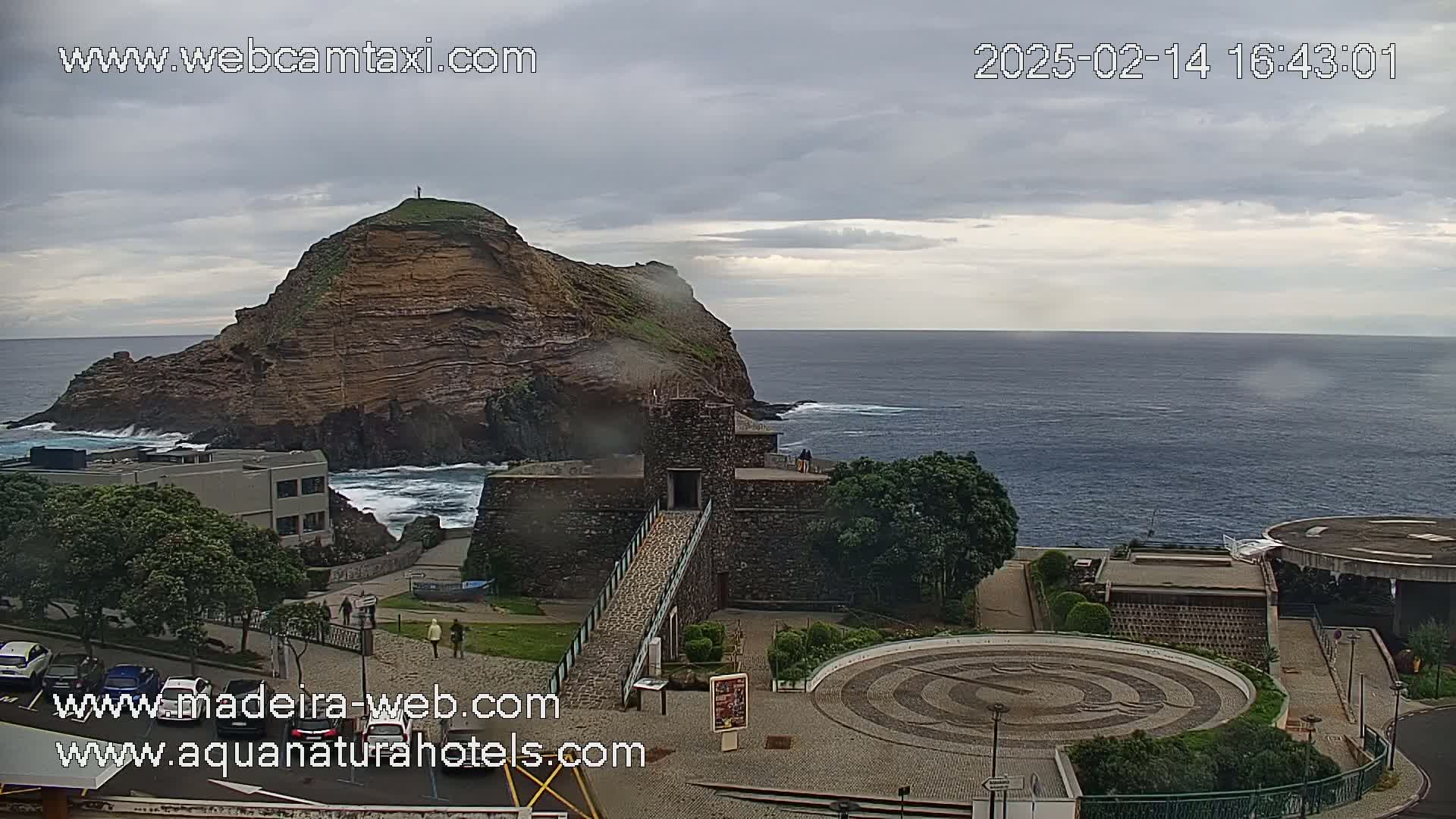 A rocky, vegetated island sits off a coast, with a stone structure and a circular paved area visible in the foreground under an overcast sky.