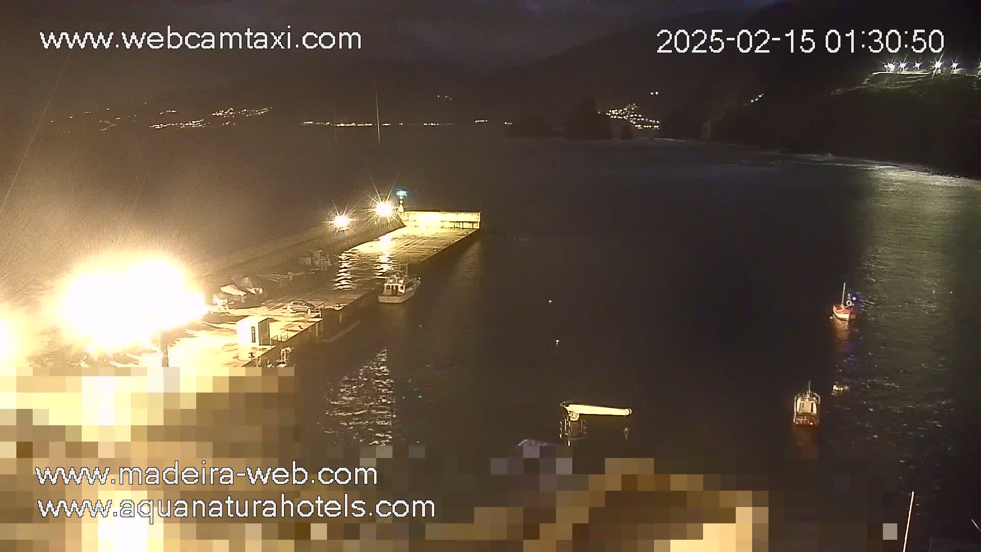 A nighttime view of a harbor with several small boats moored alongside a brightly lit dock, under a dark sky.