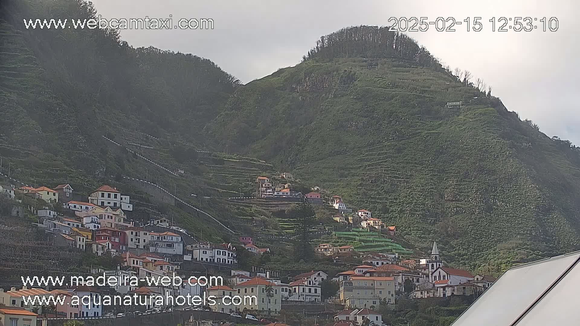 A hillside village of white and red-roofed buildings is nestled into terraced farmland on a green mountain under an overcast sky.